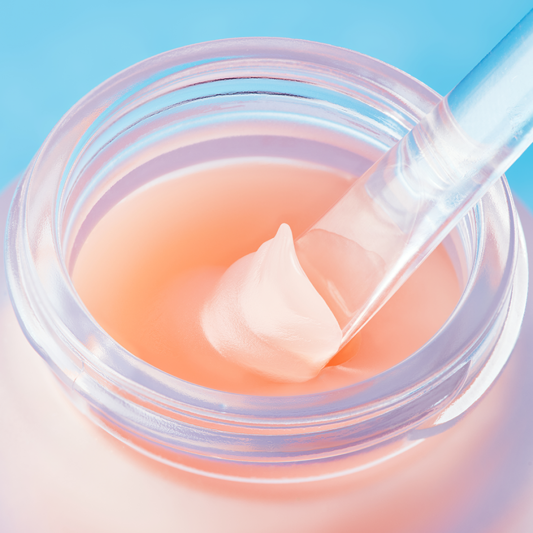 A close-up of an open jar of TOCOBO Vita Glazed Lip Mask, showing its glossy peach-colored balm texture. A clear spatula lifts a smooth scoop of the lip mask, highlighting its creamy, hydrating consistency against a soft blue background.