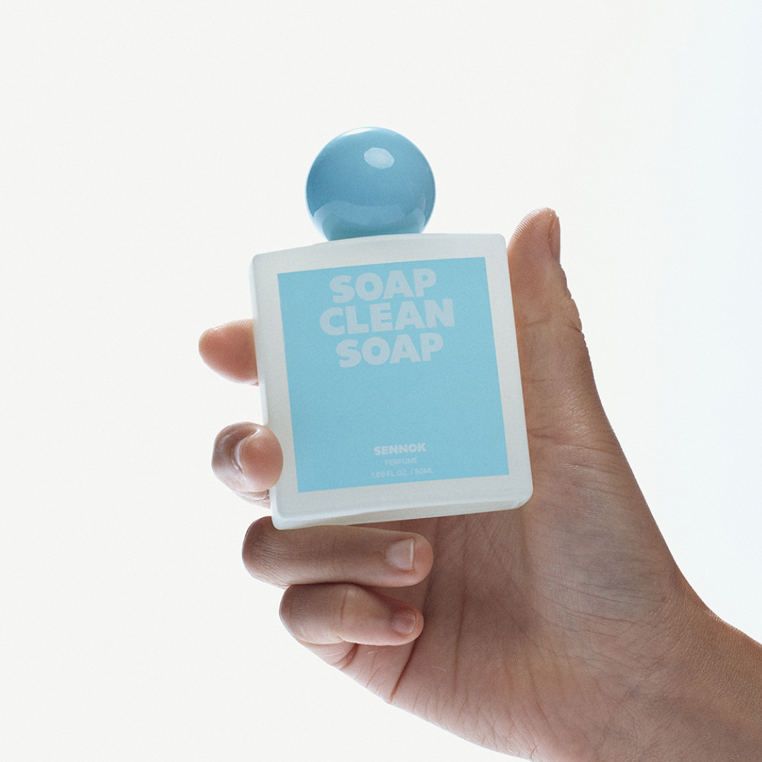 明るくミニマルな背景の中、ライトブルーのラベルと丸いブルーキャップが特徴の SENNOK「Soap Clean Soap」香水を手がそっと持ち上げているクローズアップ。