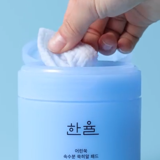 バズっている韓国コスメ:ハンユル ヤング ヨモギ ウォーター パッドの容器からパッドを取り出しているシーンで、淡いブルーの円筒型ボトルと水分を含んだ白いパッドが写っている。清潔感のある背景が、みずみずしくやさしい使い心地を表現している。