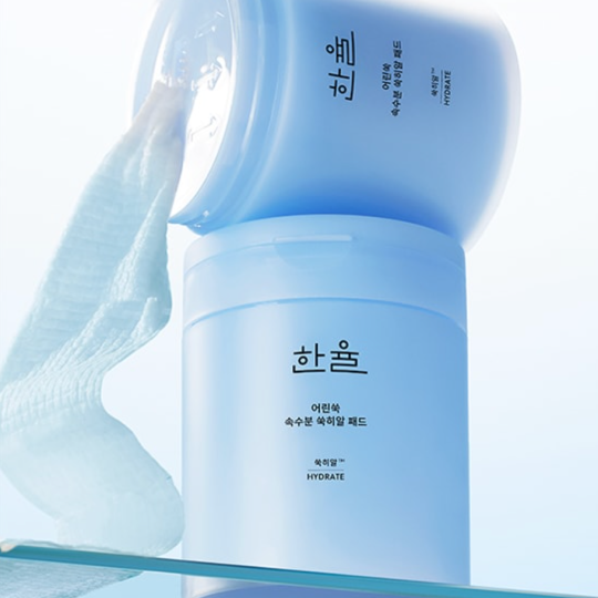 バズっている韓国コスメ:ハンユル ヤング ヨモギ ウォーター パッドの容器が開いた状態で傾けられ、中からしっとりとしたパッドが垂れ下がっているプロダクトイメージ。淡いブルートーンのミニマルなデザインが、鎮静とうるおいケアの印象を与える。