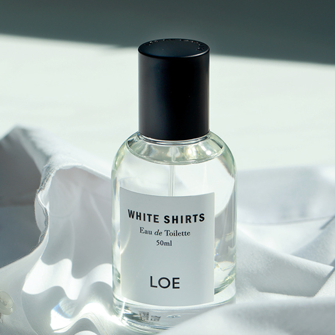 透明のガラスボトルに入った LOE の「White Shirts」オードトワレが、柔らかく折り重なった白い布の上に置かれ、自然光に照らされて清潔でフレッシュな雰囲気を演出している。