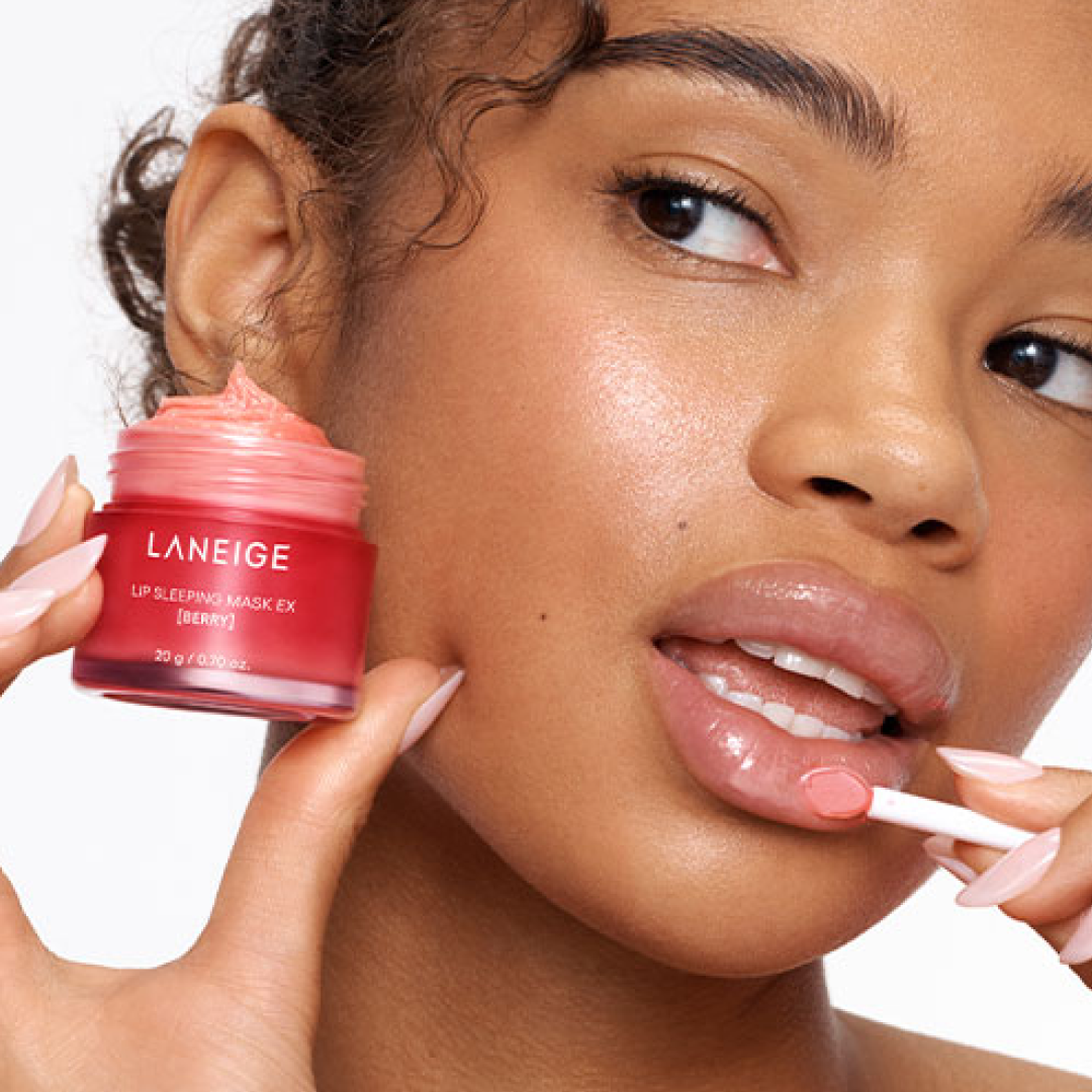 A woman applies the LANEIGE Lip Sleeping Mask EX in Berry using a small spatula. She holds the open jar close to her face, showcasing the pink, glossy balm texture. Her lips appear hydrated and shiny, highlighting the mask’s moisturizing effect.