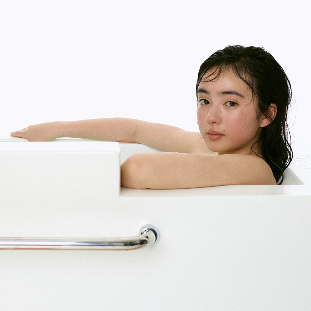 濡れた髪の若い女性が白いバスタブに寄りかかり、縁に腕を置きながらカメラに穏やかな表情を向けている。背景は明るくミニマル。