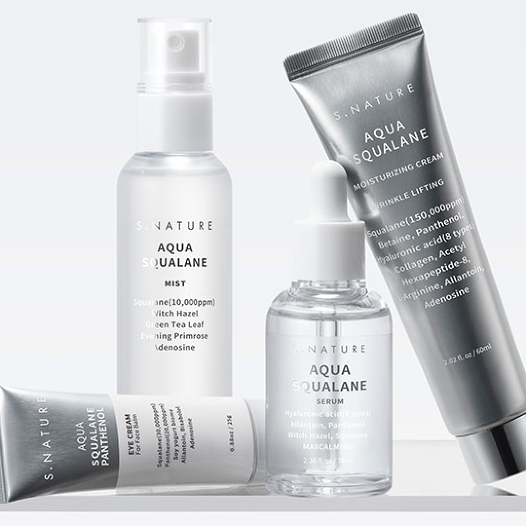 S.NATURE の Aqua Squalane ライン（ミスト、セラム、アイクリーム、保湿クリーム）が、ホワイトとシルバーを基調とした背景の上にセットで並べられている。ガラスとメタリックのパッケージが、水分補給・保湿のイメージを引き立てている。