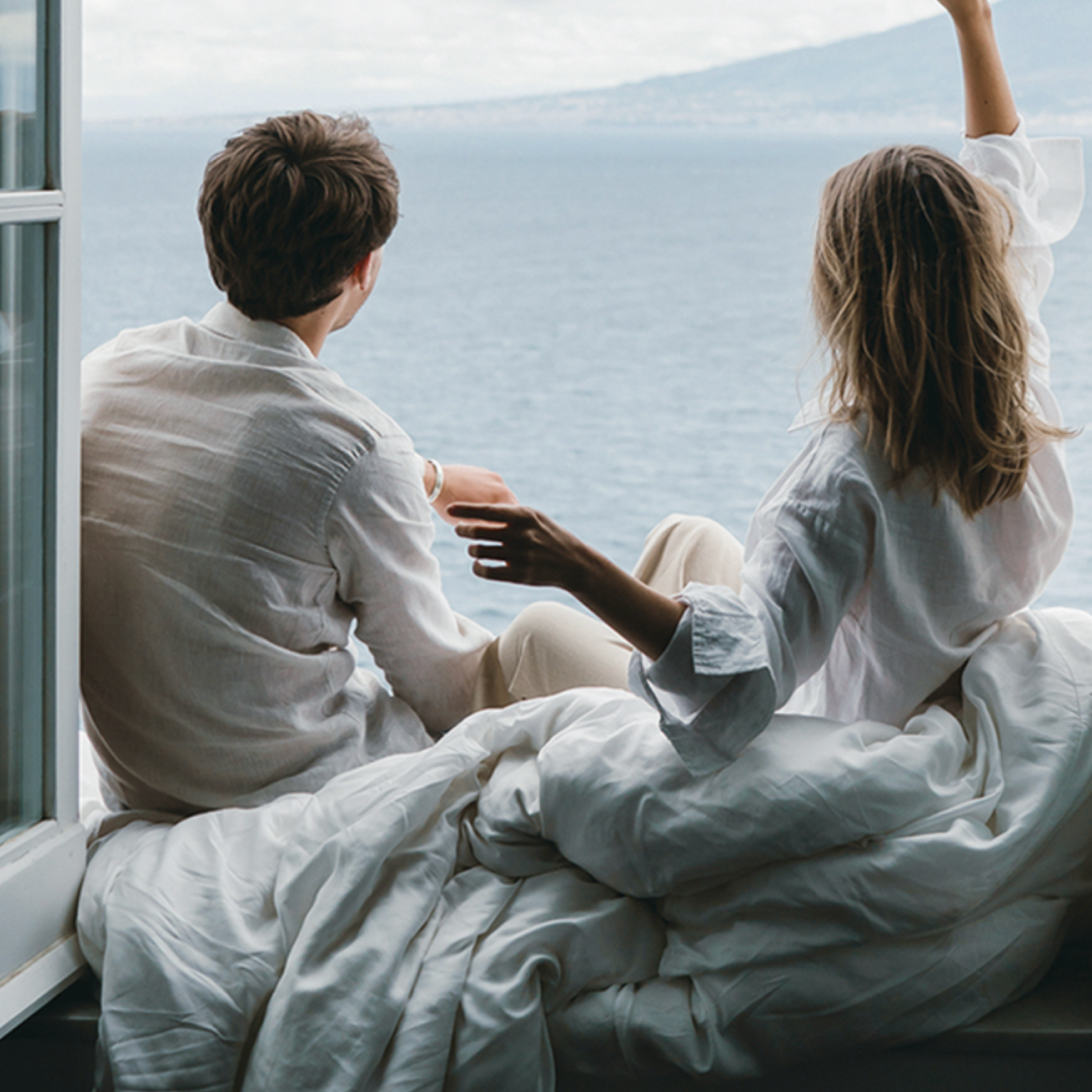 A couple dressed in light linen shirts sits on a bed by an open window, looking out at the ocean while wrapped in soft white bedding.