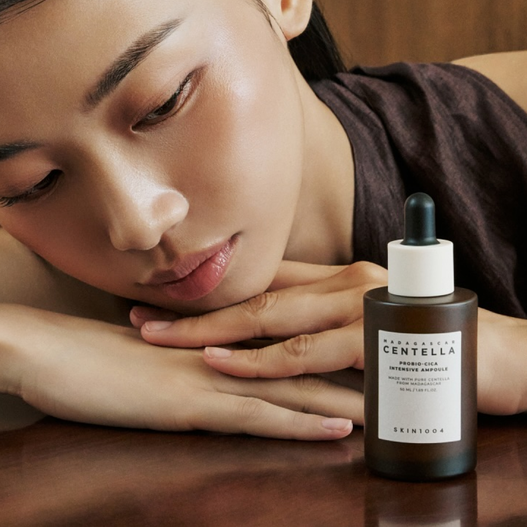 A woman rests her head and arms on a table beside a brown glass bottle of SKIN1004’s Madagascar Centella Intensive Ampoule. Her dewy, calm skin highlights the soothing and hydrating image of the product in this soft, natural composition.