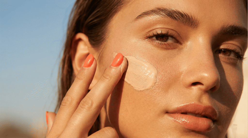 A close-up of a woman applying a smooth layer of Korean tinted sunscreen to her cheek under golden hour sunlight, demonstrating natural tone-up coverage and a dewy skin finish.