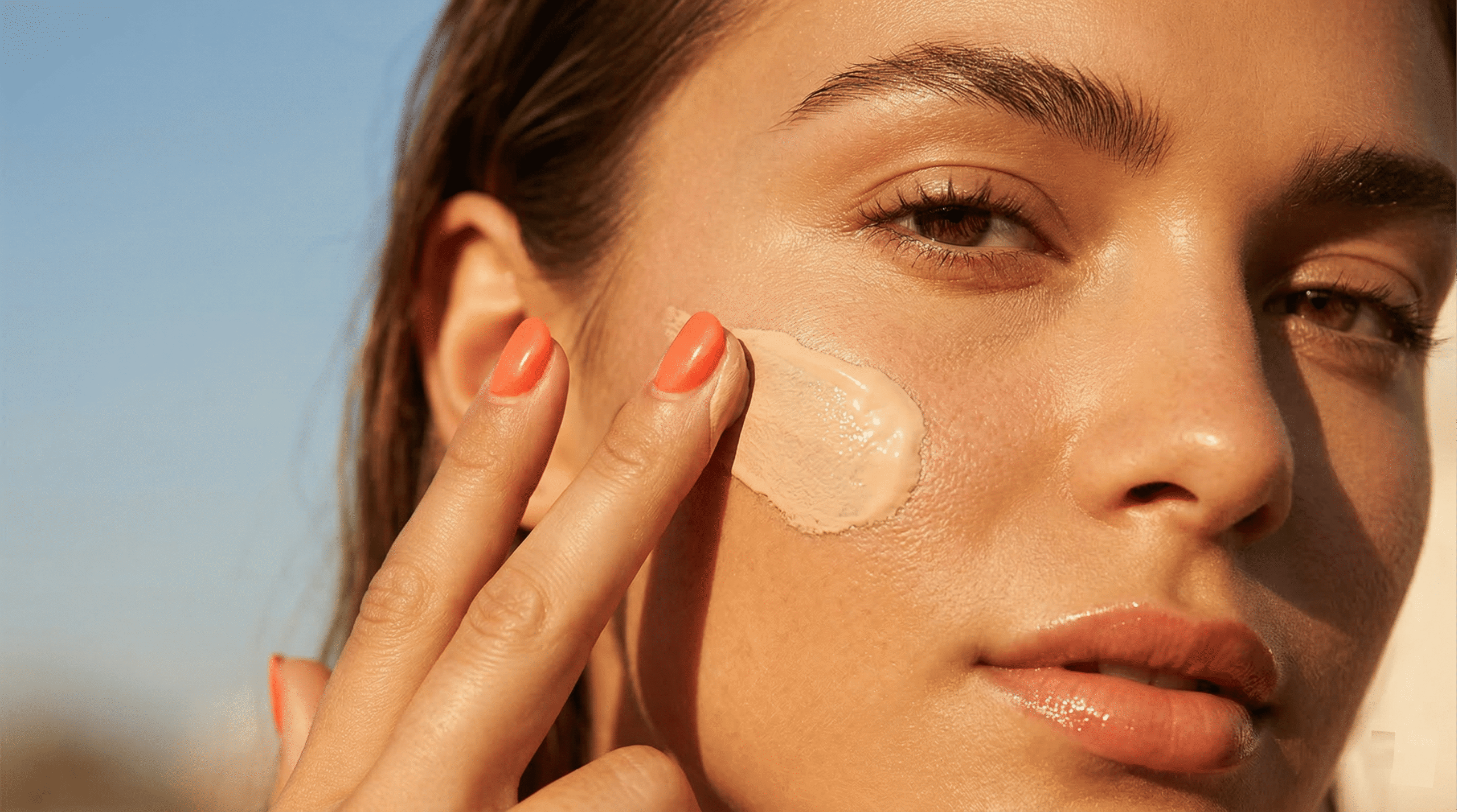 A close-up of a woman applying a smooth layer of Korean tinted sunscreen to her cheek under golden hour sunlight, demonstrating natural tone-up coverage and a dewy skin finish.