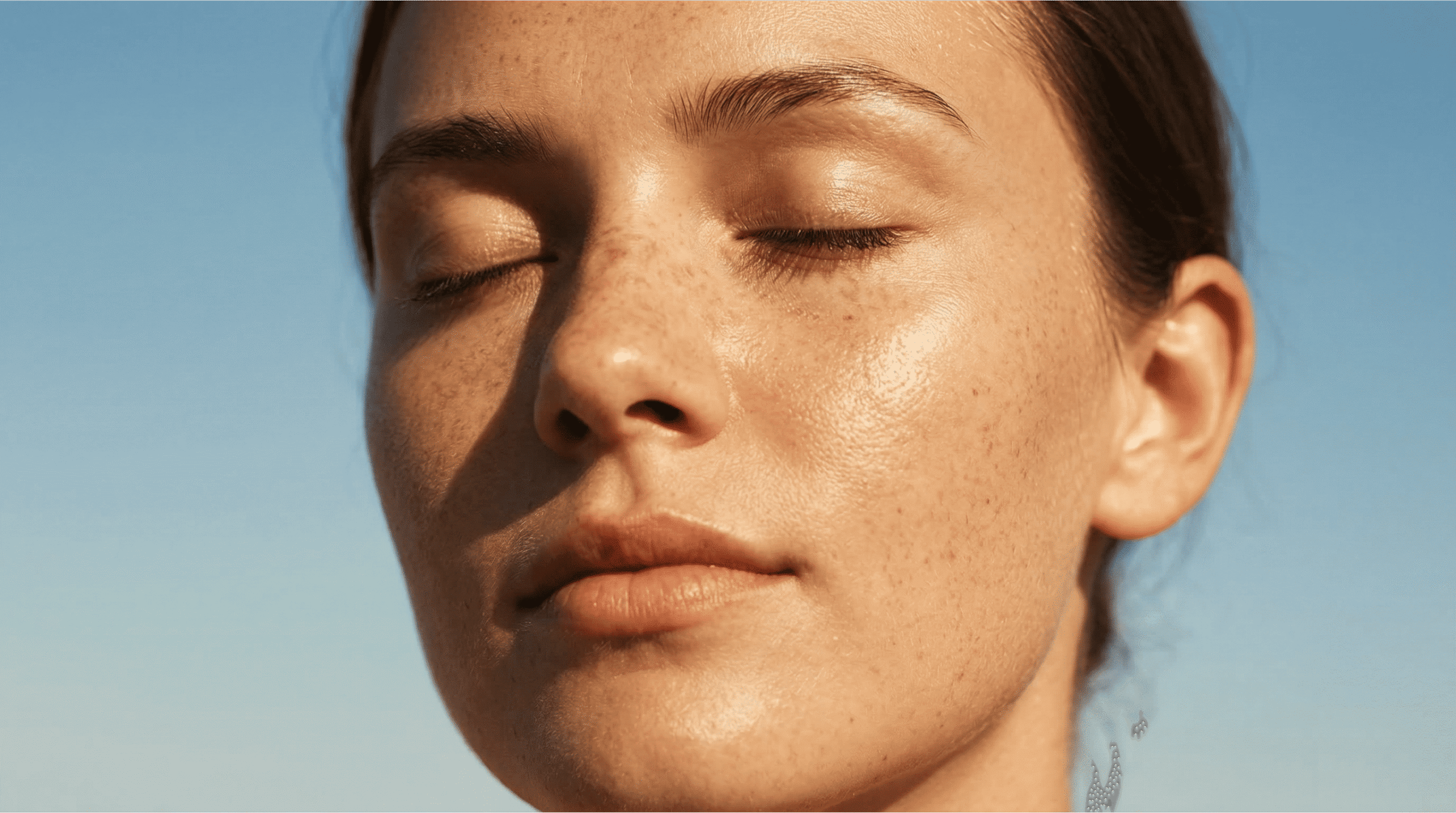 Close-up of a woman's freckled face with glowing, sun-kissed skin under direct sunlight. Her eyes are closed, showcasing a natural, dewy finish and protection provided by tinted sunscreen.