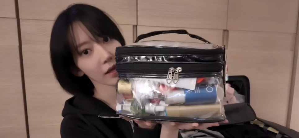 宮脇咲良がYouTube動画「What’s in my luggage」で透明ポーチに入った愛用スキンケアアイテムを紹介しているシーン。韓国コスメのボトルやアンプルが詰まったポーチを両手で持ち、カメラに見せている様子。