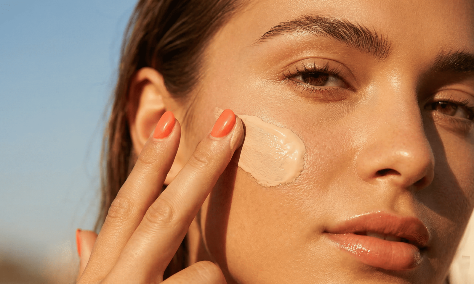A close-up of a woman applying a smooth layer of Korean tinted sunscreen to her cheek under golden hour sunlight, demonstrating natural tone-up coverage and a dewy skin finish.