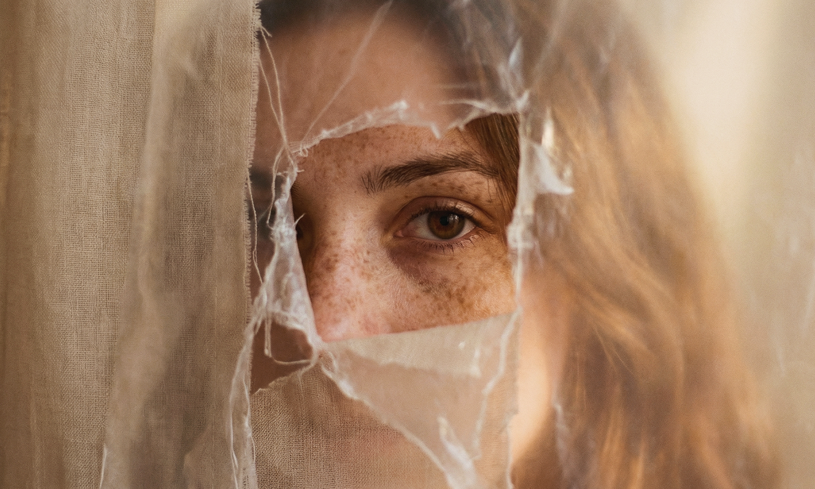 Close-up portrait of a woman’s eye partially revealed through torn beige fabric, highlighting natural skin texture in warm soft lighting.